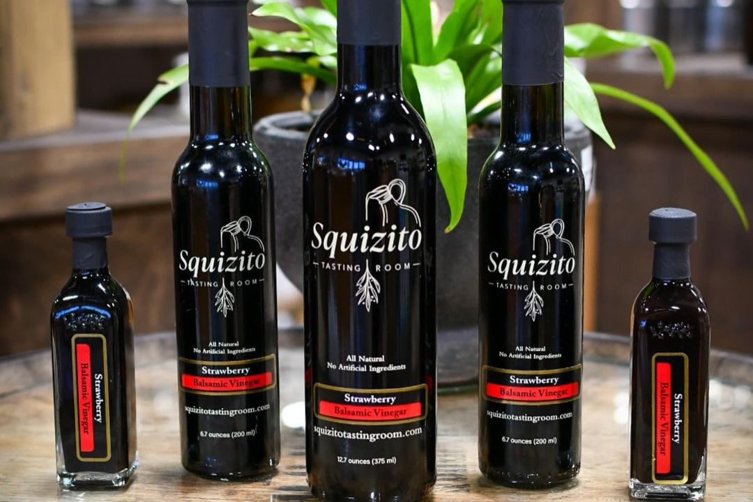 Squizito tasting room's strawberry balsamic vinegar product lineup displayed on a rustic wood table, showcasing the elegant branding, all-natural ingredients, and variety of bottle sizes. The sleek black bottles with refined labels emphasize the premium quality and unique flavors of squizito’s offerings