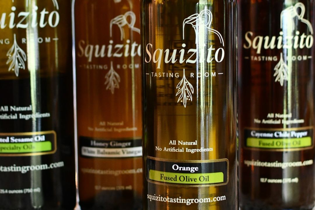 A close-up of squizito tasting room's artisanal olive oil and balsamic vinegar bottles, with the "orange fused olive oil" bottle in sharp focus. The labels highlight the brand’s commitment to all-natural, additive-free ingredients