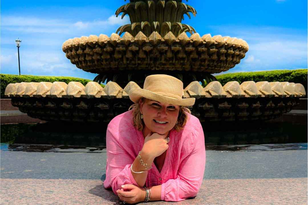 Kimberlee menchel, leaning casually by charleston's iconic pineapple fountain, embodies warmth and approachability. With a natural smile and relaxed posture, she captures the essence of professionalism with a personal touch, making her an ideal partner for clients seeking expertise infused with genuine personality.